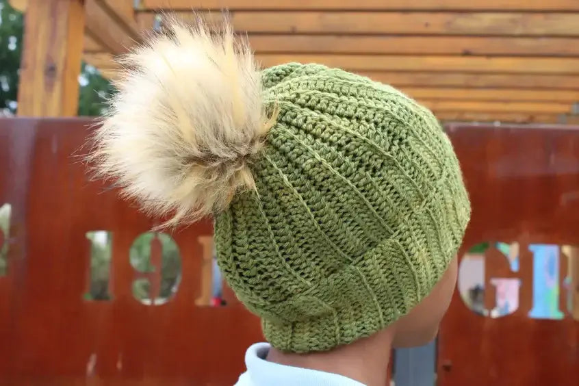 Person wearing a green knit hat with a large, fluffy pom-pom on top, standing outdoors. The wooden structure in the background and a rust-colored metal panel with cut-out numbers are visible.