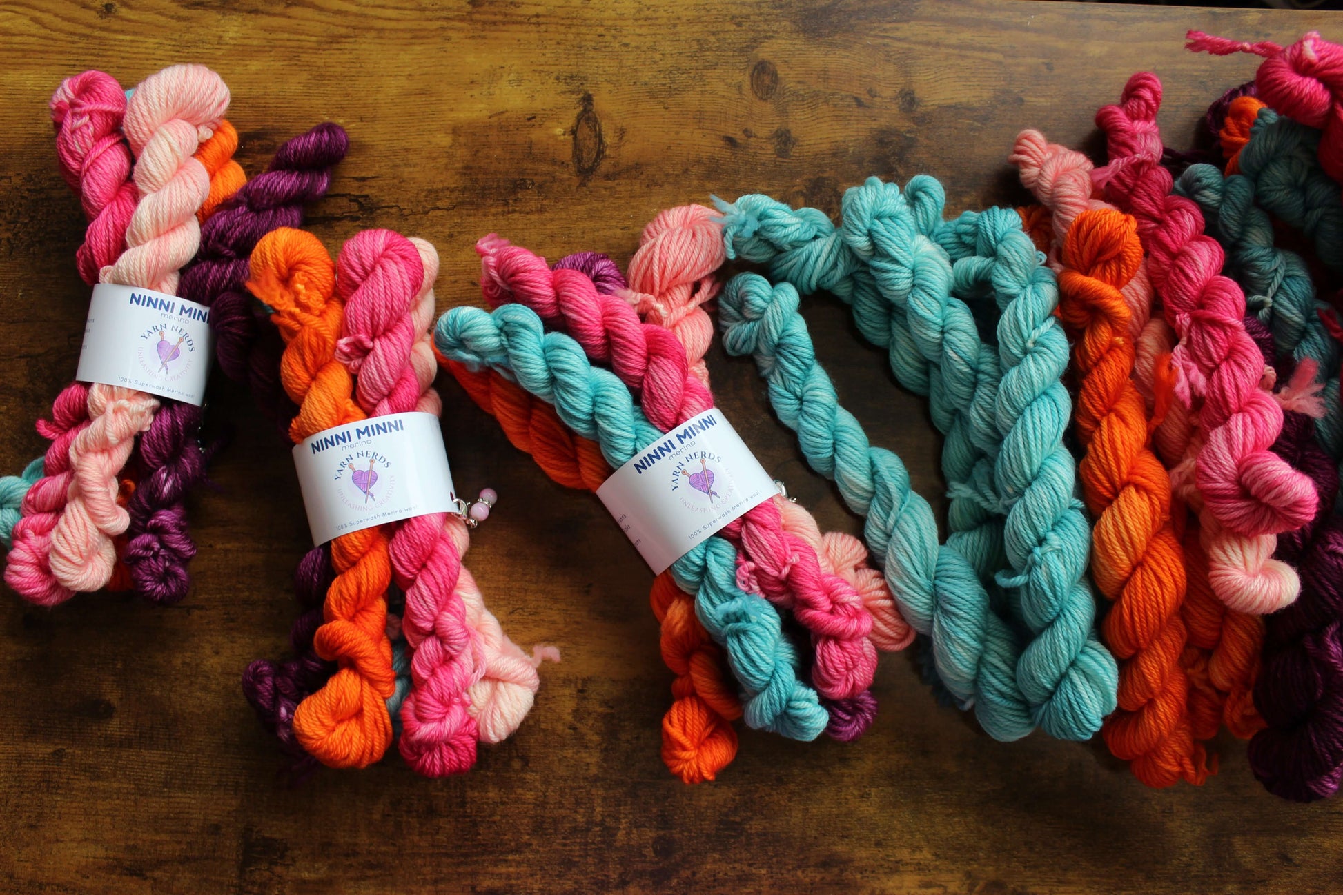 Colorful skeins of hand-dyed yarn in shades of pink, orange, purple, and teal are spread out on a wooden table. Some have labels reading SKINNY MINNIE and display a small sheep logo, showcasing Ninni Mini Merino Yarn made from superwash Merino wool.