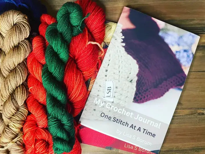 A crochet journal titled My Crochet Journal: One Stitch At A Time by Lisa S Borum is nestled beside a vibrant yarn stash, featuring skeins in beige, red, green, and orange hues on a wooden surface. Perfect for documenting your cherished crochet projects.