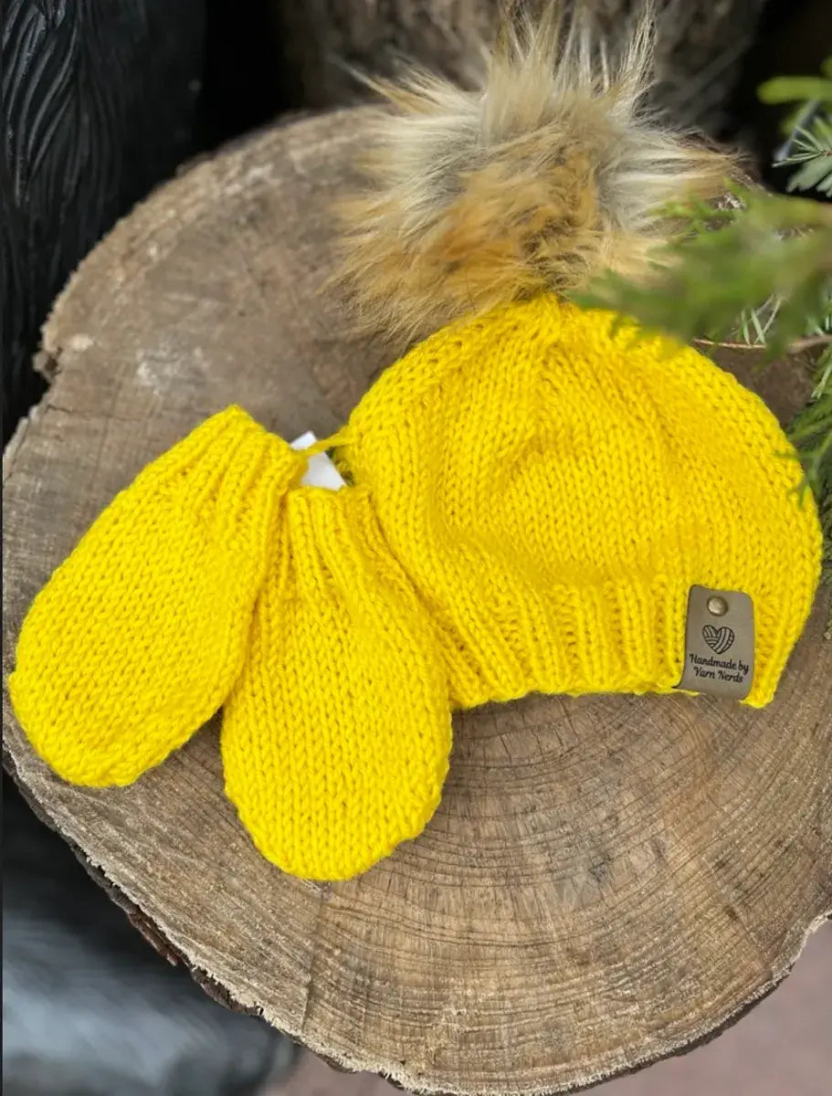 Yellow knitted Sunshine Beanie with a faux fur pompom and matching mitten set on a wooden stump. The hat features a small brown tag, complementing the cozy duo. A sprig of greenery is partially visible to the right.