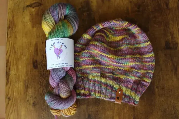 A multicolored skein of Bella Merino yarn labeled yarn nerds and a cozy Calypso Beanie Knit Pattern hat are laid out on a wooden surface. The vibrant yarn hues, including shades of purple, green, yellow, and pink, bring life to the hats design. A small leather tag adorns the hat.
