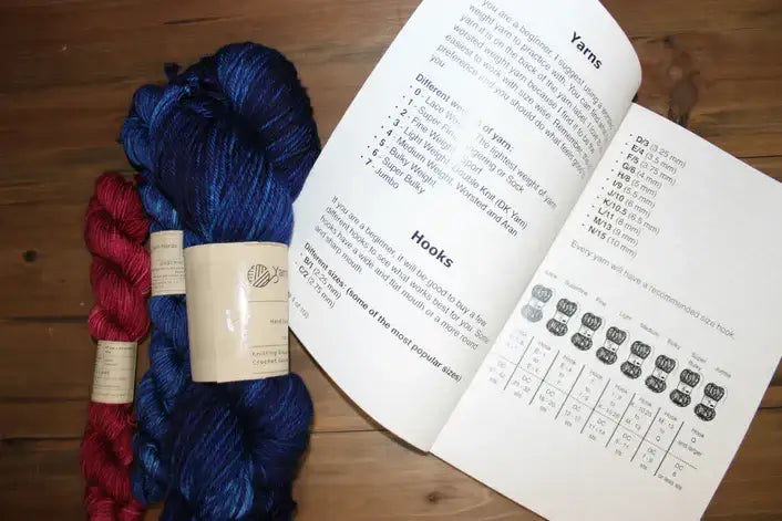 Skeins of red and blue yarn are placed on a wooden surface next to an open booklet, which resembles a crochet journal. It contains information about yarn types and crochet hooks, with illustrations and charts visible on the right page, perfect for planning future crochet projects.