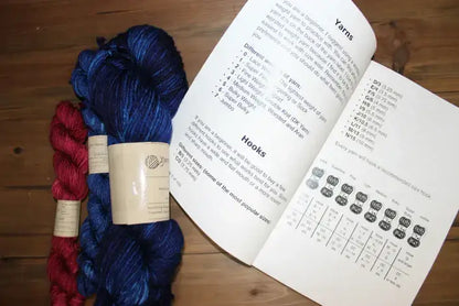 Skeins of red and blue yarn are placed on a wooden surface next to an open booklet, which resembles a crochet journal. It contains information about yarn types and crochet hooks, with illustrations and charts visible on the right page, perfect for planning future crochet projects.